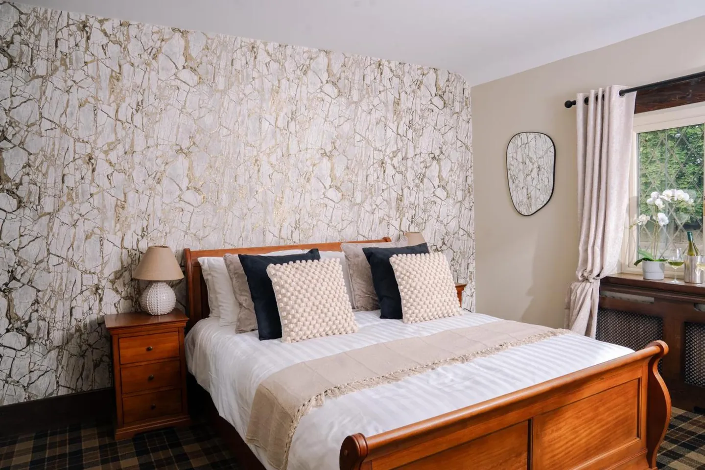 A double bed with a wooden sleigh frame against a pale stone-patterned wall, with a bedside table and a window