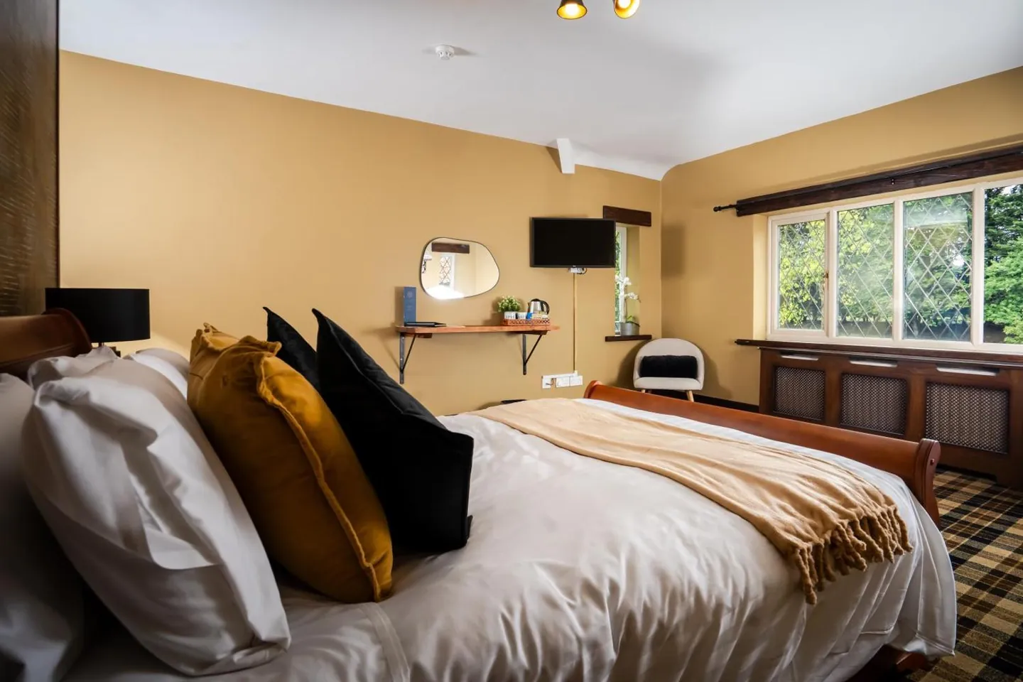 A double bed with ochre and black cushions, a wall-mounted television above a desk shelf, and a leaded window